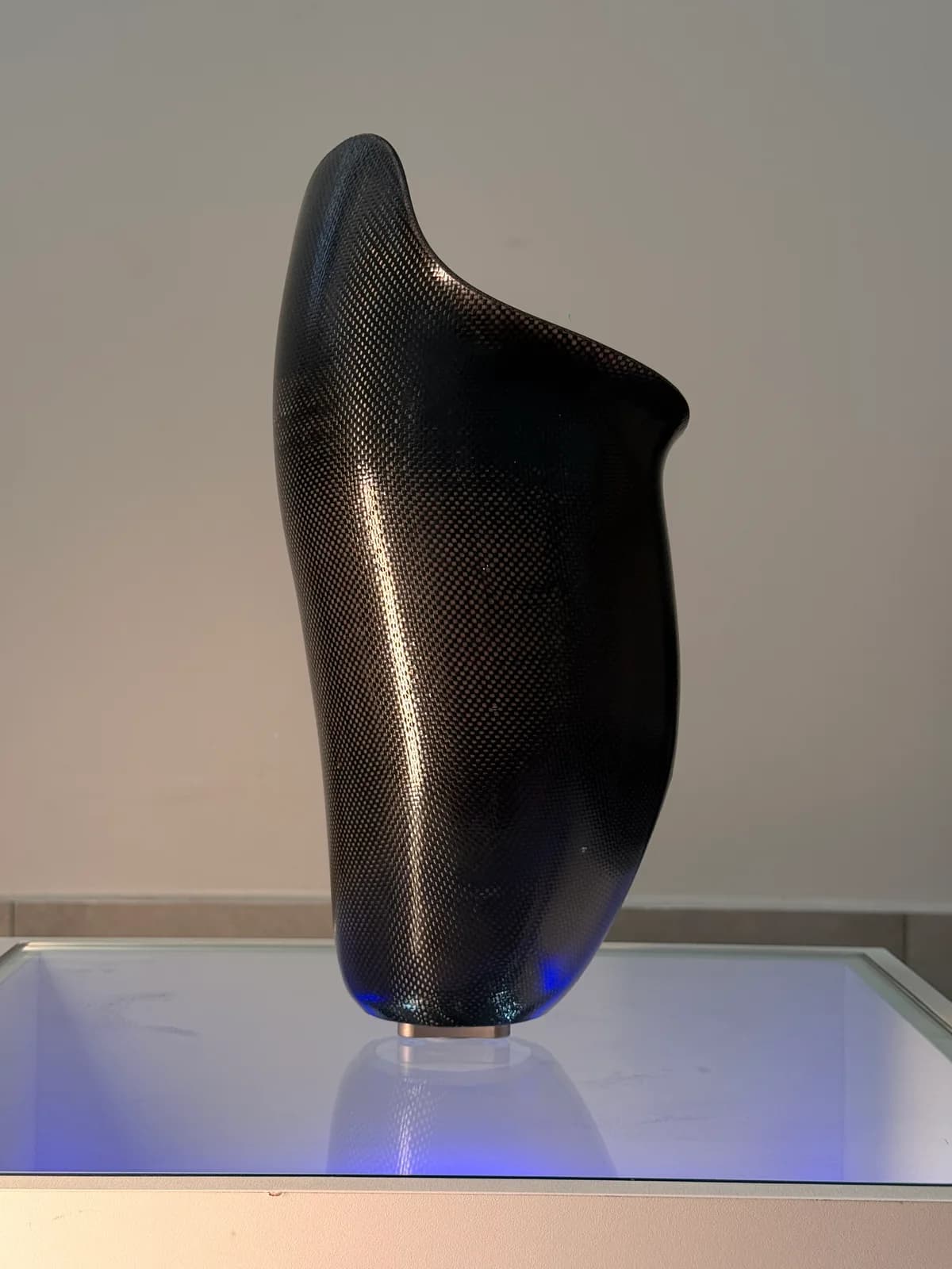 Encaixe de prótese de membro inferior em fibra de carbono, com a trama do tecido visível no acabamento, exposto sobre uma base iluminada